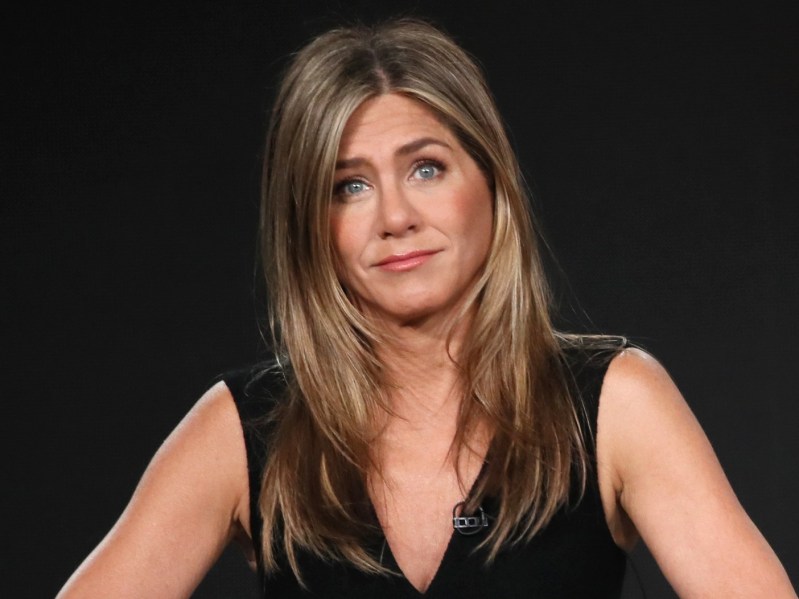 Jennifer Aniston sits while wearing a black dress. There is a pitch black backdrop behind her