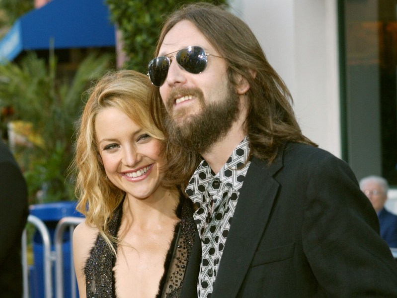 Kate Hudson (L) smiles in a sequined gold dress and Chris Robinson smiling in black blazer and sunglasses