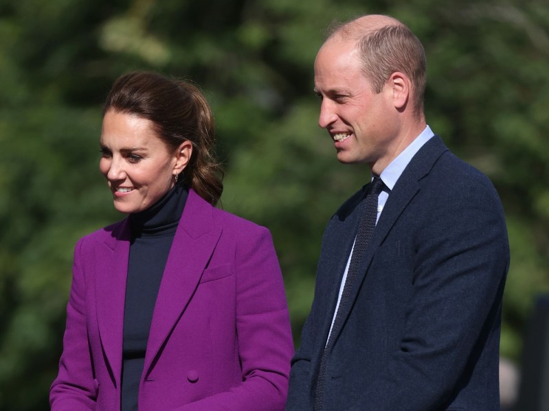 Kate Middleton (L) wearing black top with bright purple blazer, standing outside next to Prince William, who is dressed in a classic navy blue suit and tie