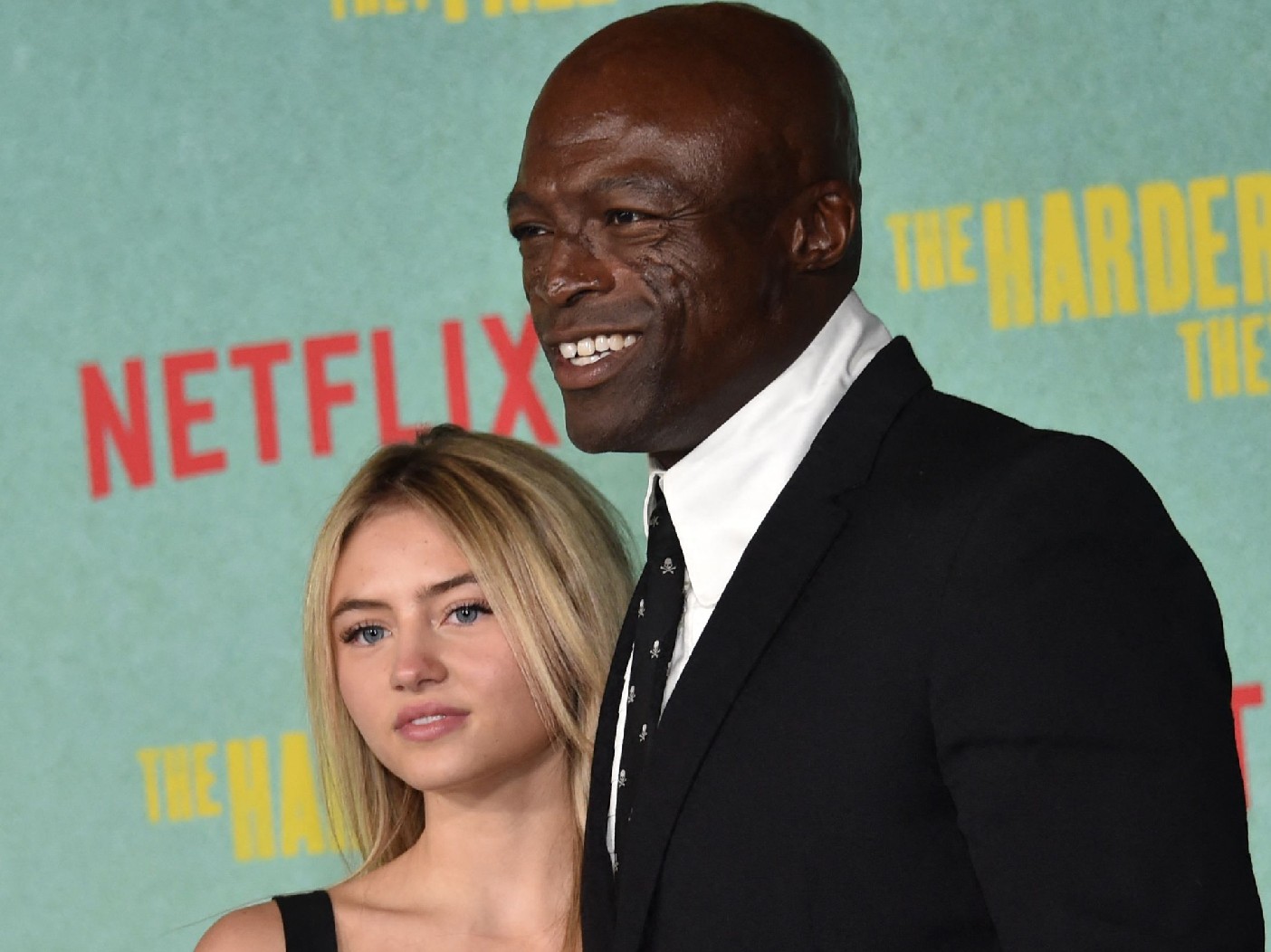 Closeup of Leni Klum (L) looking ahead wearing black strapped dress. On her right is Seal, also looking ahead, dressed in a black suit and tie