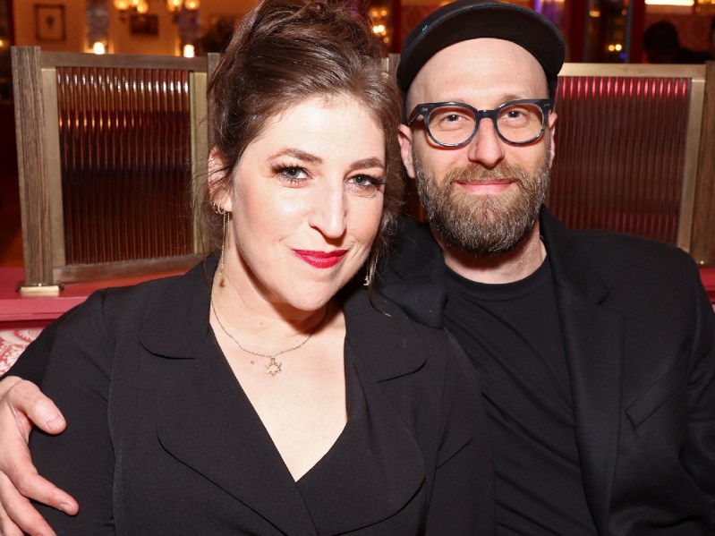 Mayim Bialik, in a black blazer, sits with Jonathan Cohen, also dressed in black, indoors