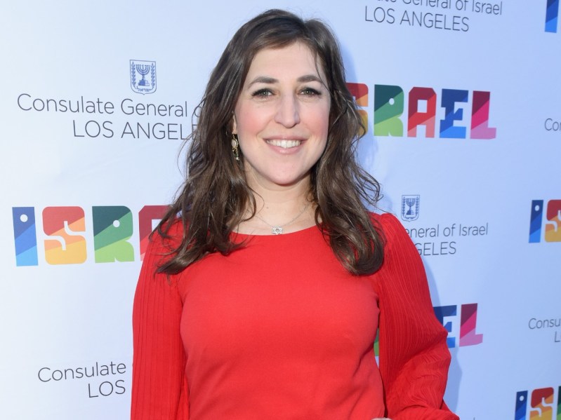 Mayim Bialik smiles with a hand on her hip. She is wearing a long-sleeved orange dress