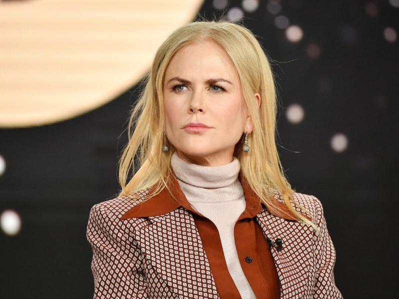 Nicole Kidman wears a sienna colored outfit over a cream colored sweater against a dark background