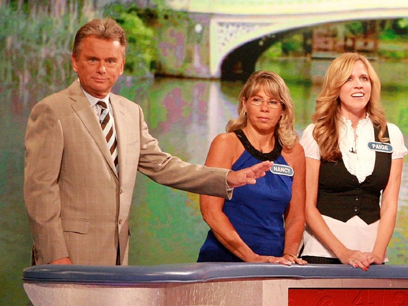 Pat Sajak wears a tan suit on the set of Wheel Of Fortune as he stands near several contestants