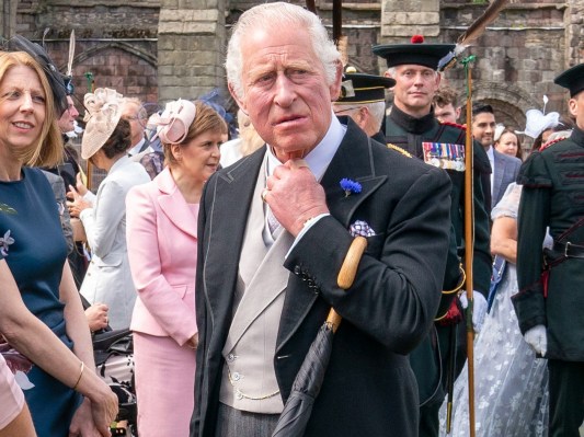 Prince Charles looks off into the distance. He is standing in the middle of a crowd outside.