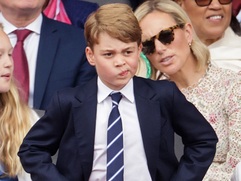 Prince George shands with his hand on his hips with a quizzical look on his face. He is wearing a navy blue blazer with a white button down shirt and navy and white striped tie