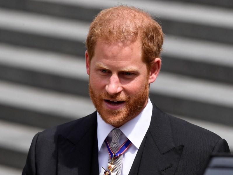 Prince Harry wears a medallion around his neck over a white dress shirt with a black blazer and patterned tie