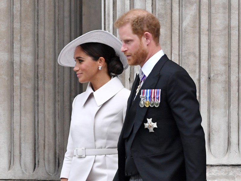 Meghan Markle (L) wearing white blazer and white hat, walking with Prince Harry, who is dressed in a classic suit ensemble