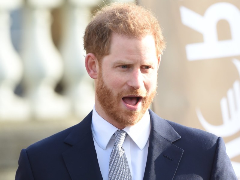 Prince Harry stands with his mouth wide open. He is wearing a white dress shirt with a navy blue blazer and light gray tie