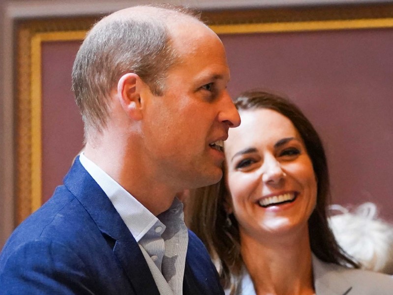 Prince William wears a blue suit and stands with his wife Kate Middleton