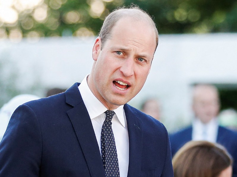 Prince William appears in mid-speech outside while wearing a navy blue blazer over a white dress shirt and matching tie