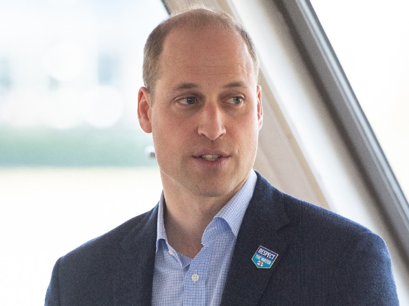 Prince William looks off into the distance. He is wearing a suit and blue dress shirt
