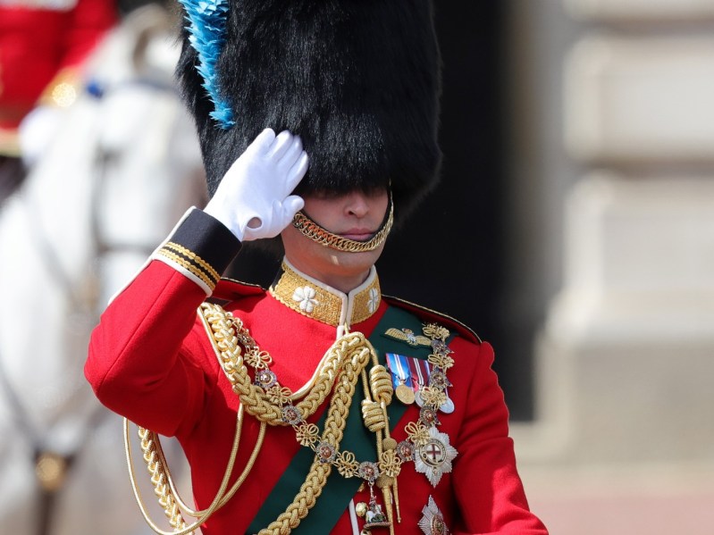 Prince William rides horseback in a red suit jacket with green and gold sash. He has his hand to his forehead in a salute