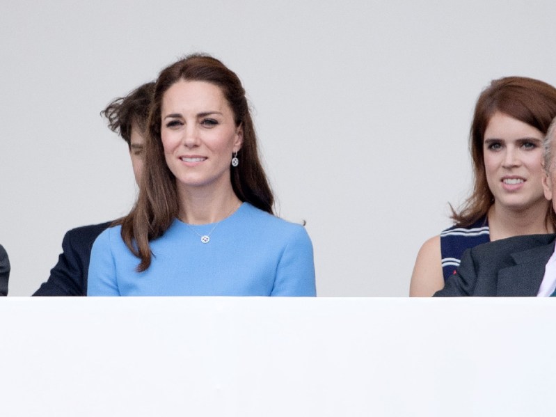 Kate Middleton wears a blue dress and sits slightly in front of Princess Eugenie, in a dark dress