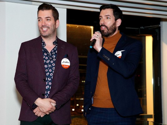 Drew and Jonathan Scott stand together in jaunty suits while one brother holds a microphone on a makeshift stage