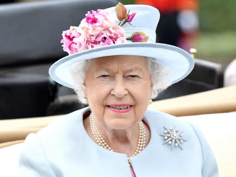 Queen Elizabeth smiles while wearing pale blue top and matching hat with pink flower details