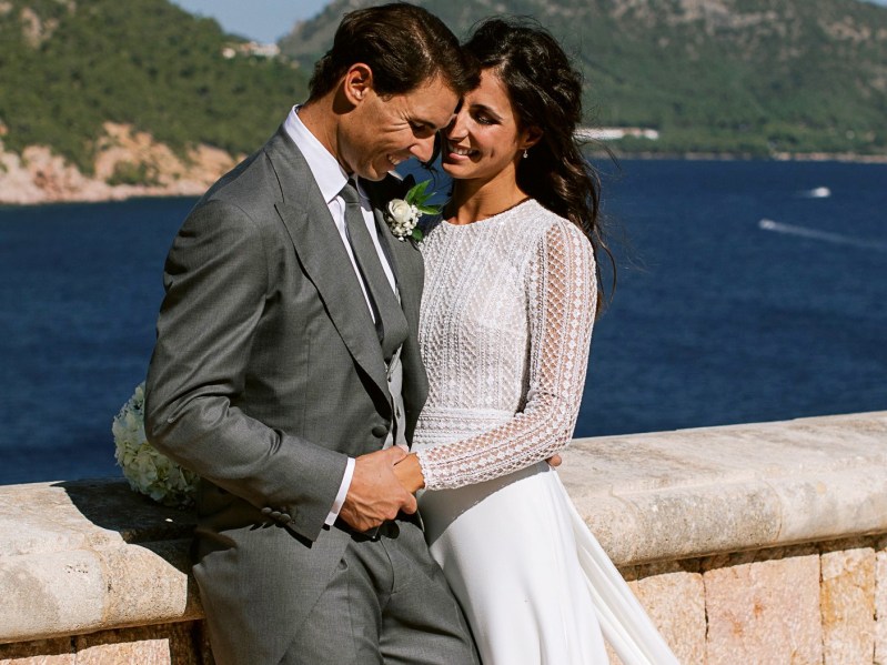 Rafael Nadal (L) in a gray suit and his wife Mery in a wedding dress. The two are standing close together and smiling at each other.