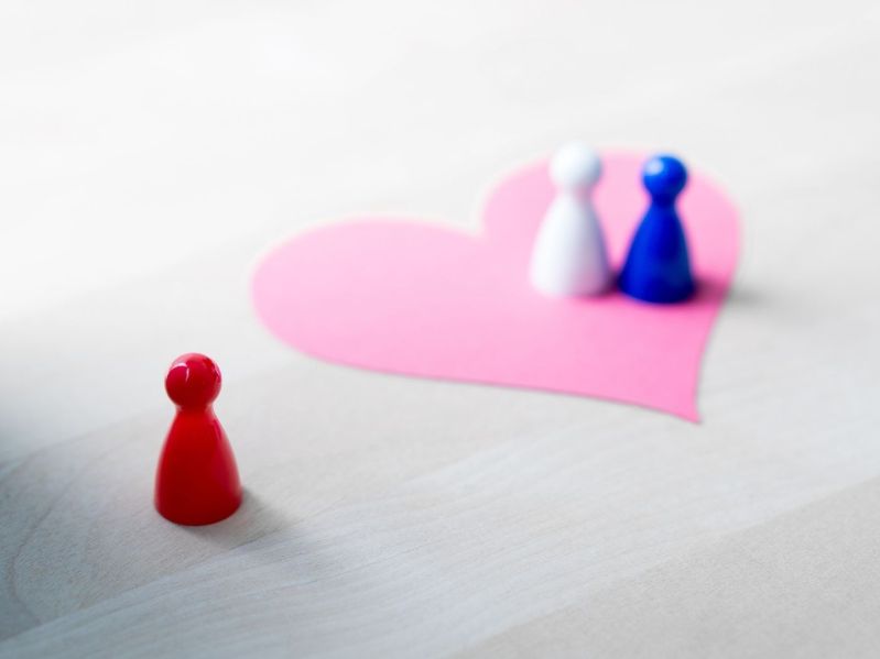 Two board game pawns atop a paper heart on table with a third pawn set apart