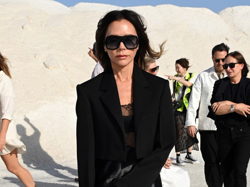 Victoria Beckham in sunglasses and an all-black outfit walking in front of a white background on a beach