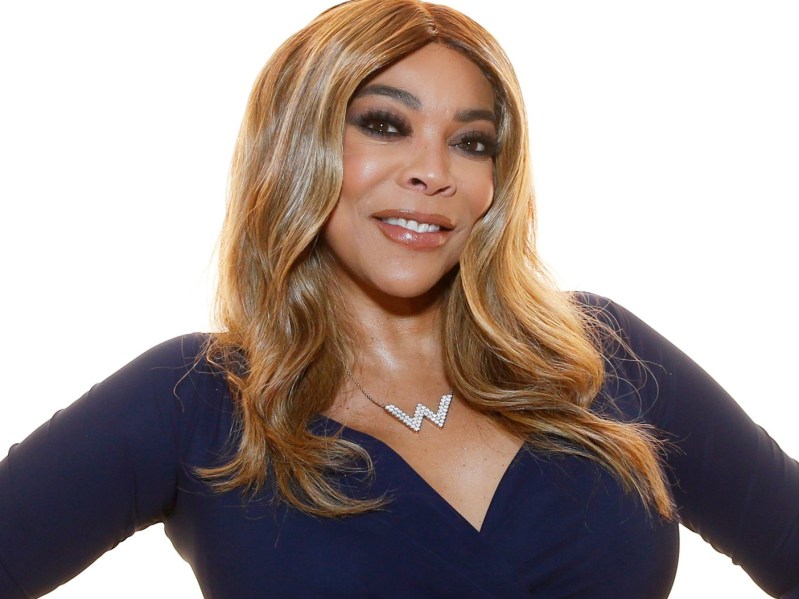 Wendy Williams wears a black dress against a white glowing background on the red carpet