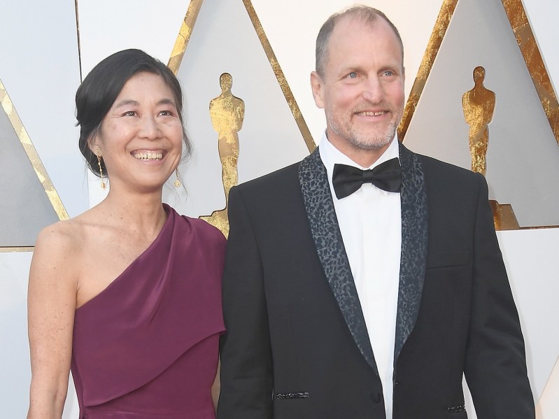 Laura Louie (L) wearing one-shoulder burgundy gown, standing next to Woody Harrelson, who is wearing a classic black suit and tie