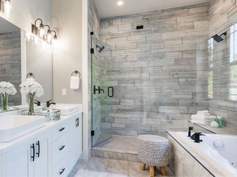 A remodeled bathroom with white double-sink vanity, gray tiled double shower with glass door and a soaking tub