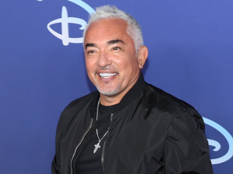 Cesar Milan smiles against blue background while wearing a black shirt with black jacket