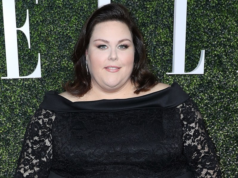 Chrissy Metz wears a black lace dress and is standing against a green bush background