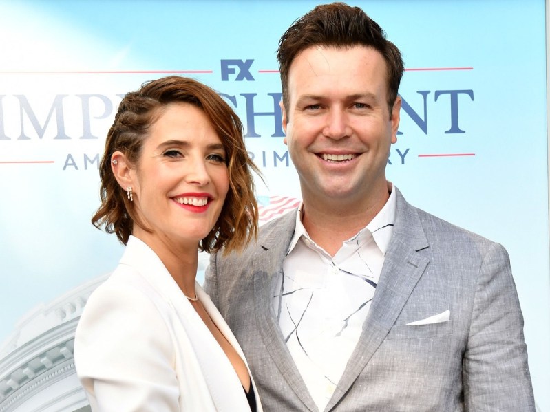 Cobie Smulders (L) wearing white pantsuit, standing next to Taram Killam, who is wearing a gray blazer over a white dress shirt