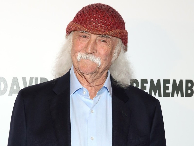 David Crosby smirks in black suit jacket over pale blue dress shirt. He is also wearing a red hat