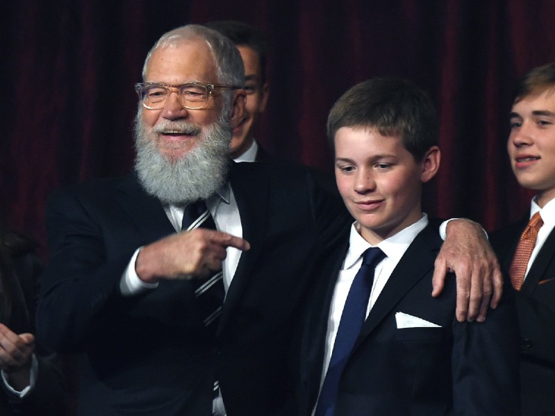 David Letterman (L) wears a classic black suit and tie and points to his son, who is standing next to him. His son is also dressed in a suit and tie