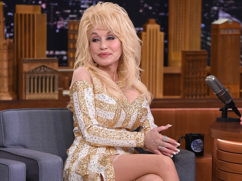 Dolly Parton smiles in white and gold dress while sitting on the stage