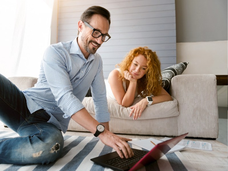 A man sits on the floor in front of a laptop as a woman lounges behind him on a couch