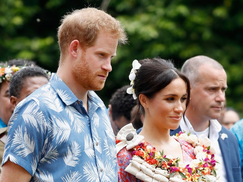 Prince Harry (L) wearing blue printed top standing next to Meghan Markle, who is wearing red patterned dress