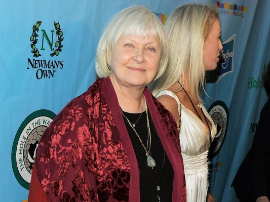 Joanne Woodward smiles against blue backdrop while wearing red jacket over black top