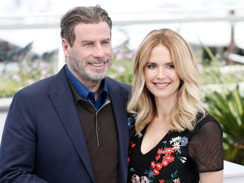 John Travolta (L) wearing navy suit jacket over black three-quarter zip sweater, standing next to Kelly Preston, who is wearing black dress with embroidered flowers