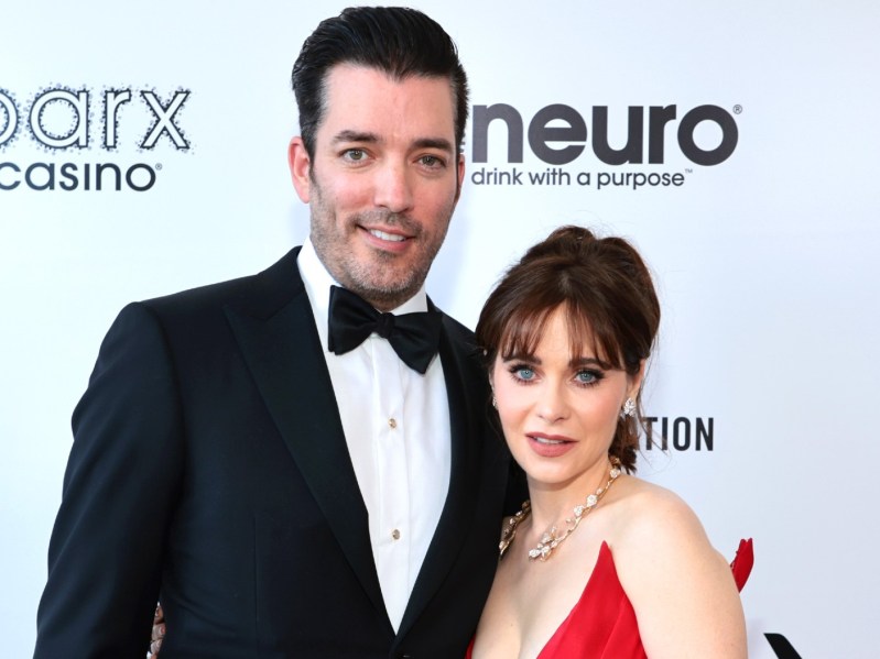 Jonathan Scott (L) in classic black suit and tie, standing next to a much shorter Zooey Deschanel, who is wearing a red gown