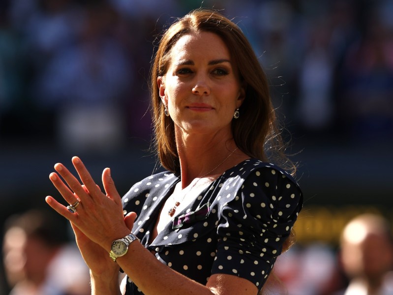 Kate Middleton looks off into the distance with her hands clasped. She is wearing a navy blue and white polka-dotted dress