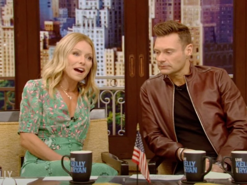 Kelly Ripa (L) speaks whily wearing a green dress, with Ryan Seacrest next to her. He is straight-faced and wearing a black top with a brown leather jacket on top