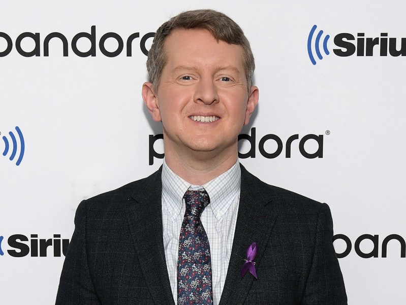 Ken Jennings smiles in classic black suit with patterned tie against white backdrop