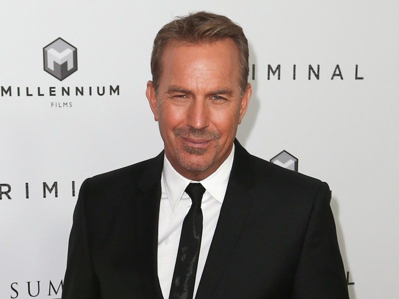 Kevin Costner smiles in classic black suit and tie against white backdrop