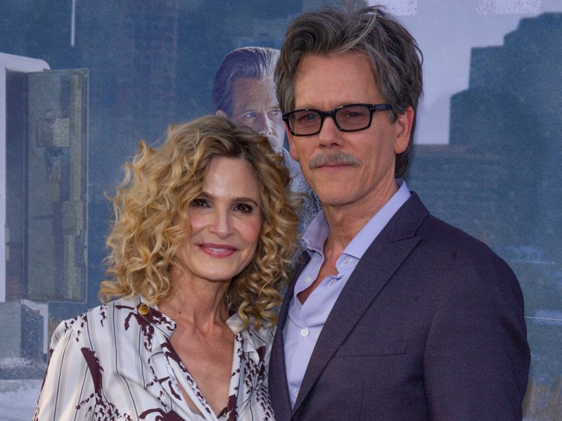 Kyra Sedgwick (L) wearing patterned silk top standing next to Kevin Bacon, who is wearing a suit and sunglasses