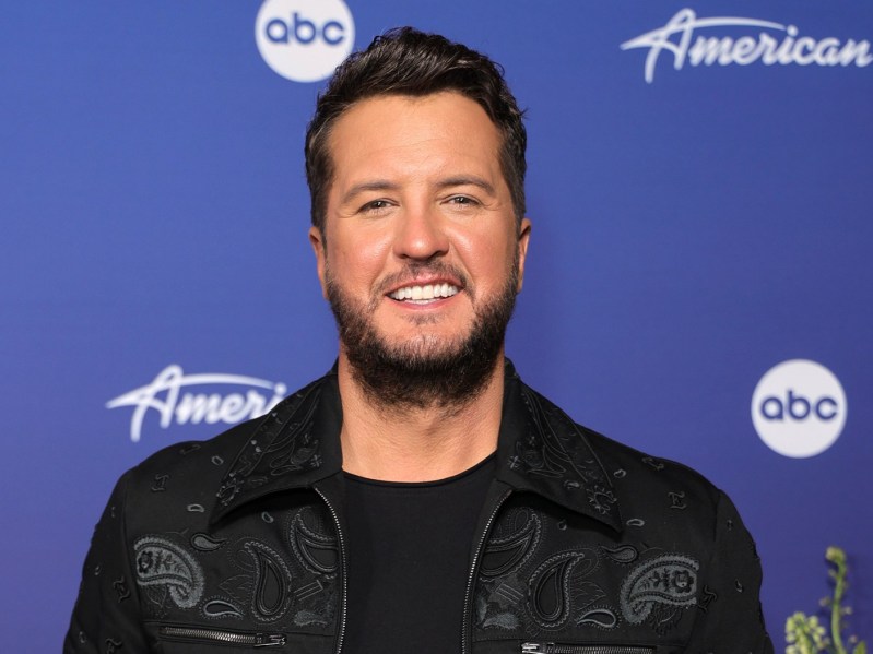 Luke Bryan smiling in black denim jacket against blue backdrop