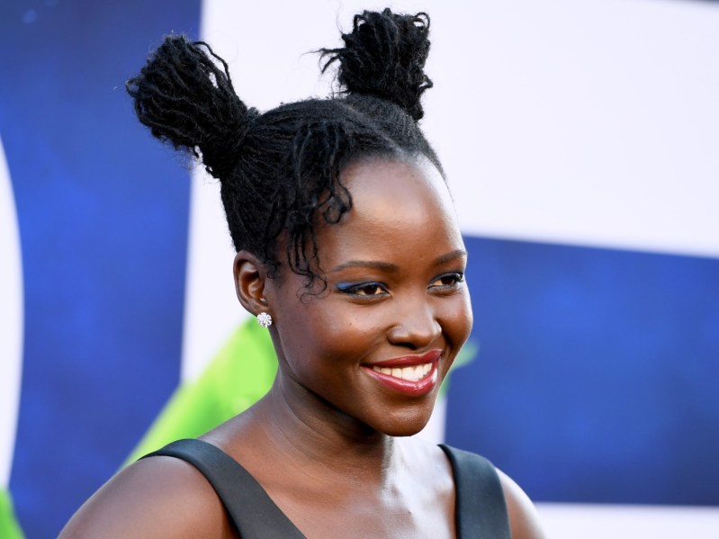 Lupita Nyong'o smiles in a closeup with her hair in two buns against a blue and green patterned backdrop