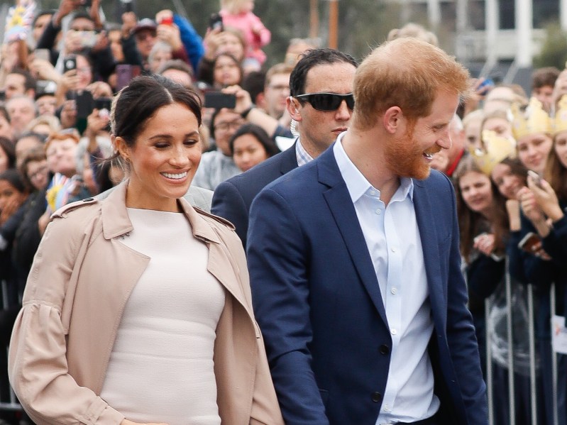 Meghan Markle (L) laughs in white dress with beige coat, walking with Prince Harry who is dressed in a navy blazer over a white dress shirt
