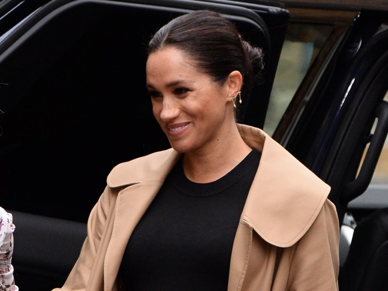 Meghan Markle smiles in black shirt with beige/tan coat