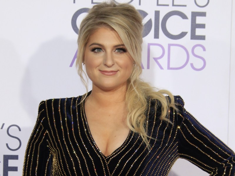 Meghan Trainor smiles, wearing a black long-sleeved dress with vertical gold pinstripes