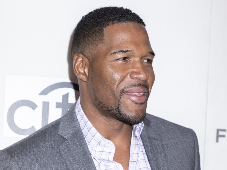 Michael Strahan smiling in side profile. He is wearing a white dress shirt and gray suit jacket