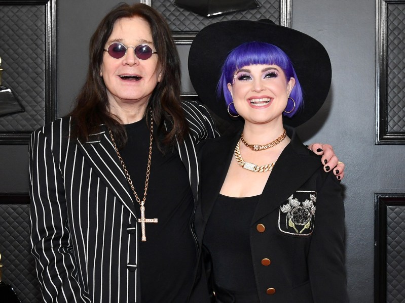 Ozzy Osbourne (L) in striped suit with cross necklace standing next to Kelly Osbourne, who has on a purple wig and black outfit