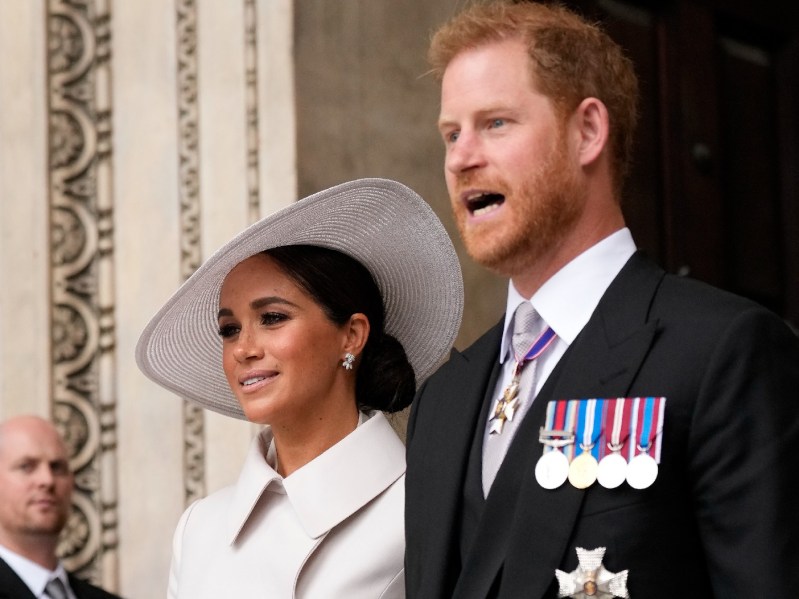 Meghan Markle, in a white coat and hat, stands with husband Prince Harry, in a dark suit jacket with medals on the lapel, outside of a church